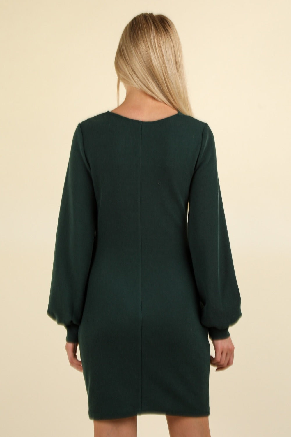 A person stands facing the camera, wearing the VERY J Ruched Detail Bodycon Mini Dress in dark green, featuring a V-neck and a wrap-style bodice. The figure-hugging silhouette accentuates their form beautifully, set against a plain, light-colored background.