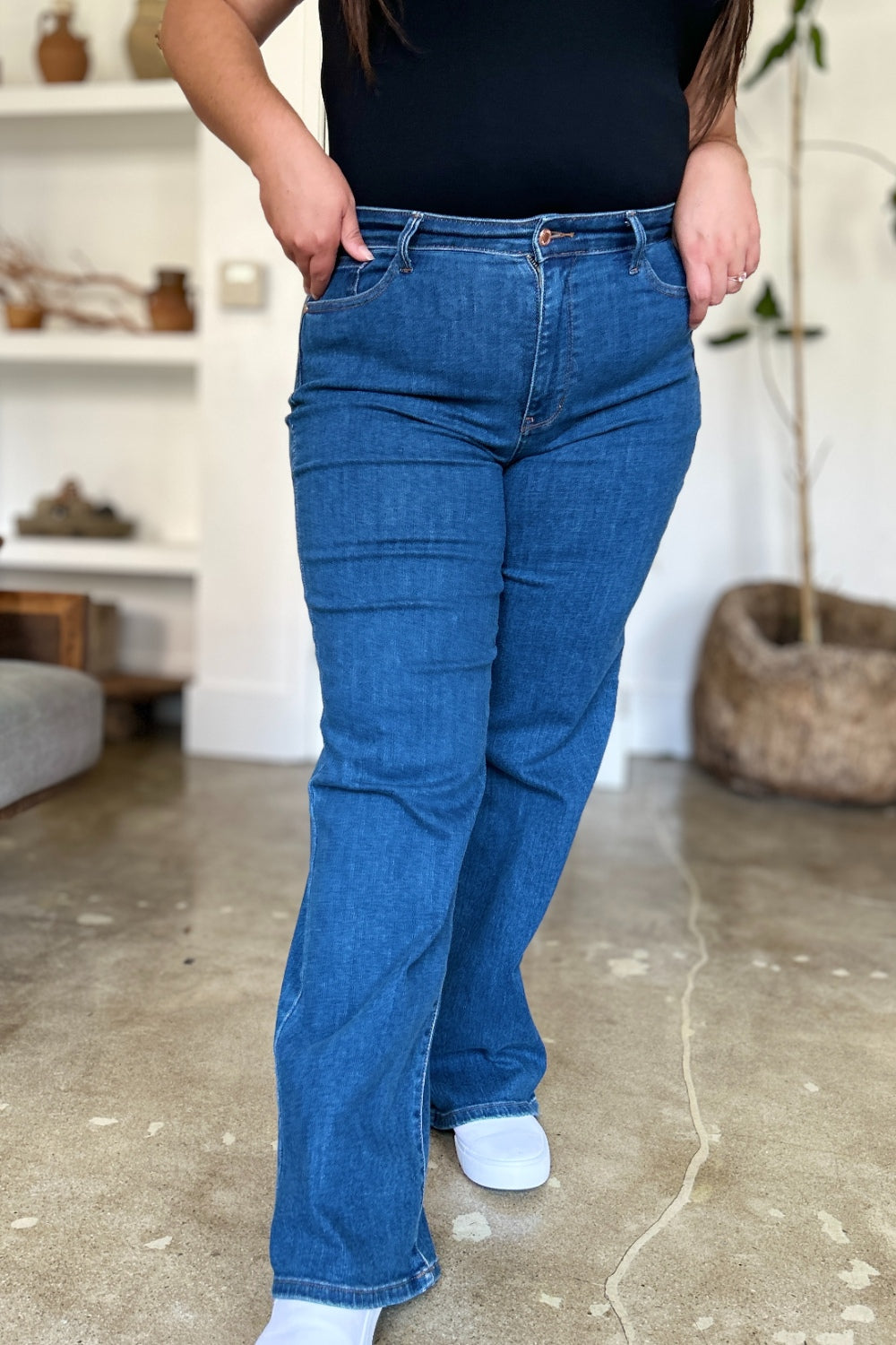 A person wearing Judy Blue Full Size High Rise Straight Jeans, a black top, and white shoes stands gracefully on a polished concrete floor.