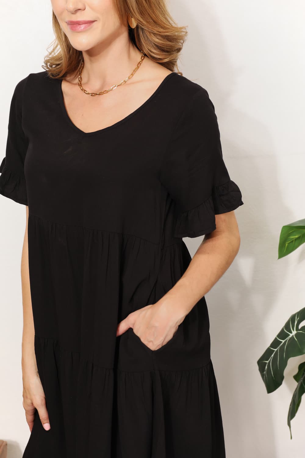 A woman stands indoors wearing the Mandy V-Neck Flounce Sleeve Tiered Dress in navy blue. She has one hand on her hip and is smiling.