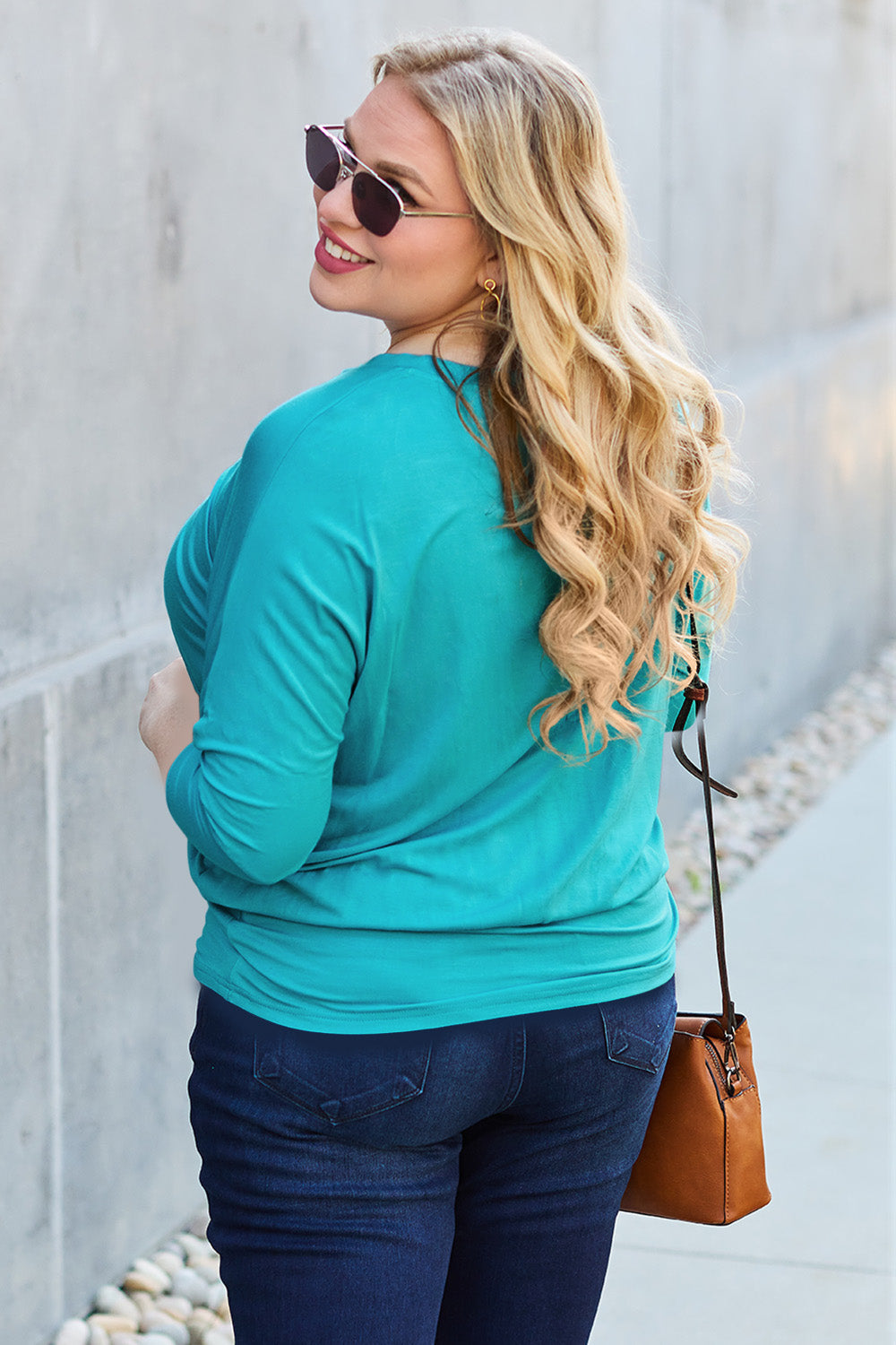 A woman in a slightly stretchy, brown Basic Bae Full Size Round Neck Batwing Sleeve top and blue jeans stands against a concrete wall, holding a brown handbag.