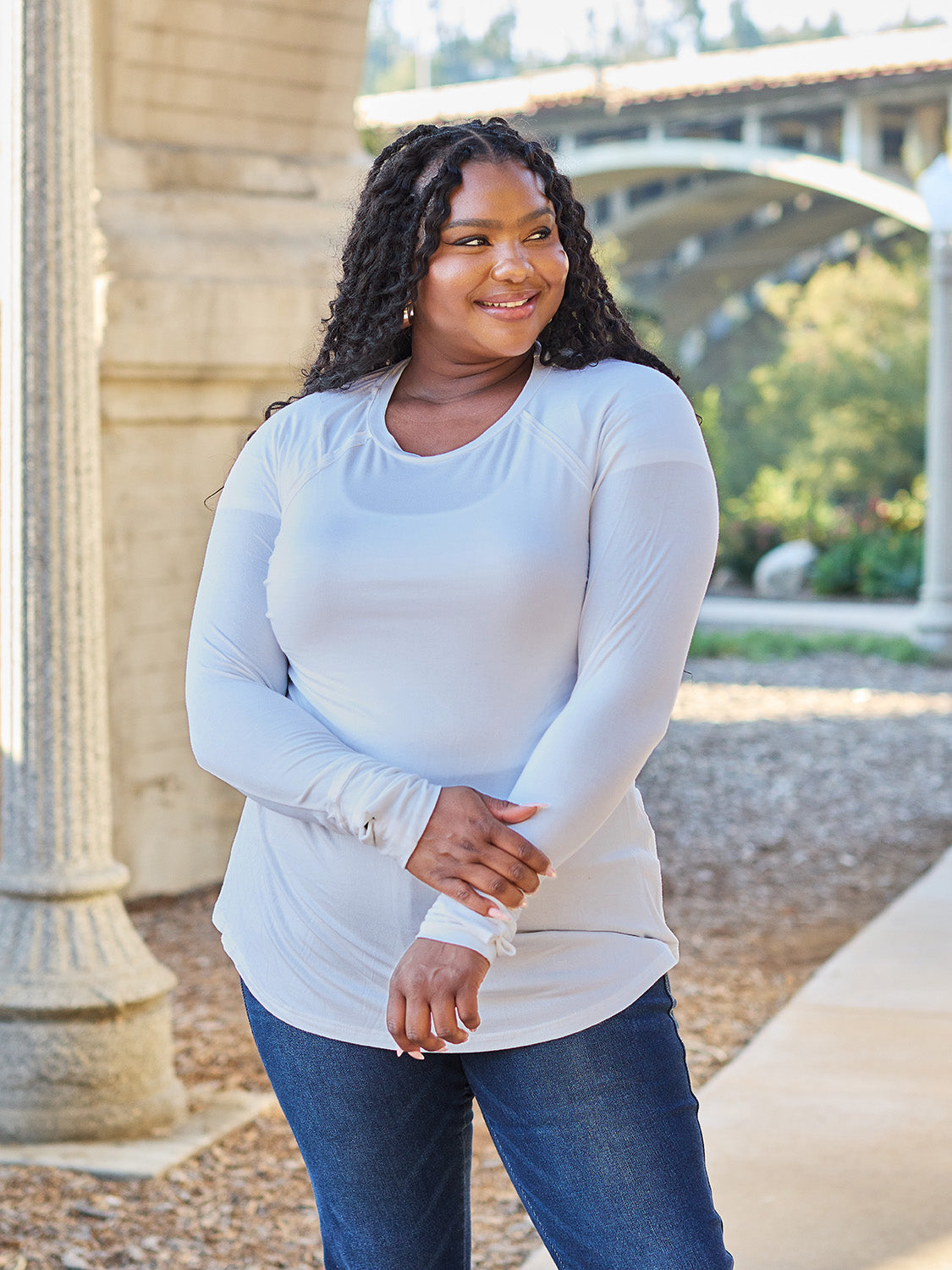 A woman wearing sunglasses and a slightly stretchy Basic Bae Full Size Round Neck Long Sleeve T-Shirt in white stands on a sidewalk with an arched structure in the background, smiling slightly.