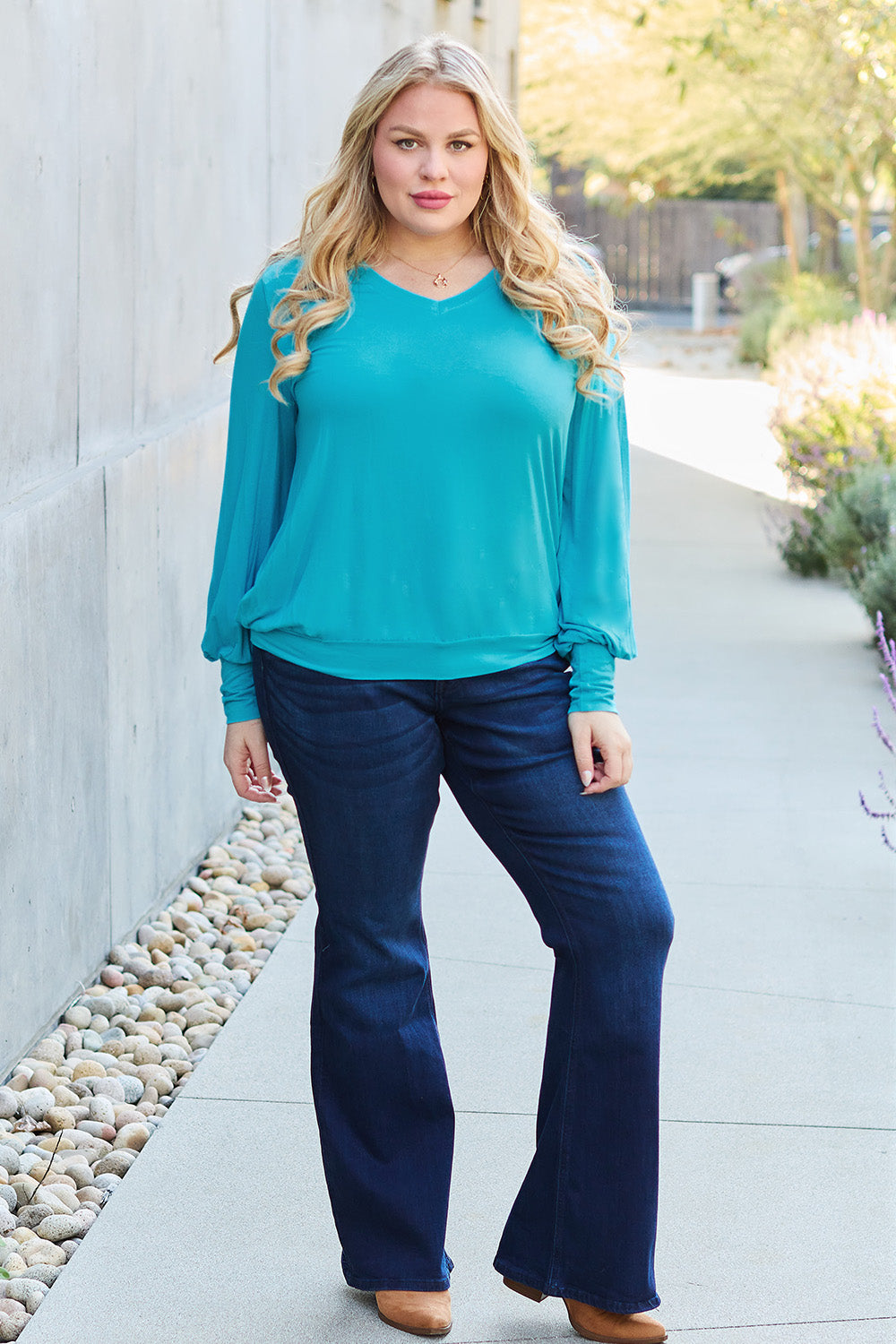 A woman with brown hair is standing against a concrete wall, wearing the Basic Bae Full Size V-Neck Lantern Sleeve Top in teal and blue basic-style jeans, smiling.