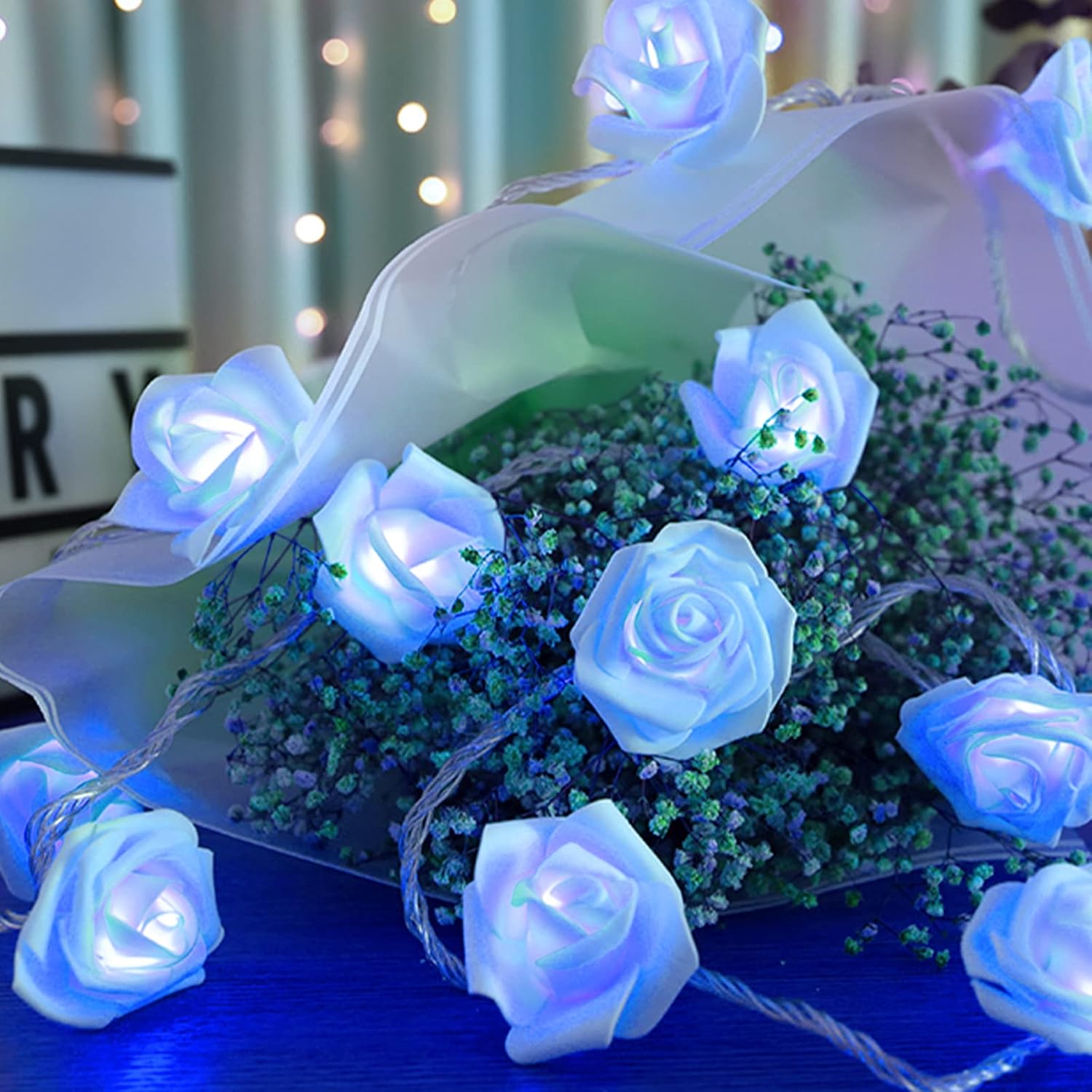 The Blue Rose Flower String Lights - 14.7ft 30 LED Fairy Lights Flower Garland Lights are elegantly draped over a stack of books and a framed photo, creating a romantic, glowing decor for any occasion, whether it's indoors or outdoors.