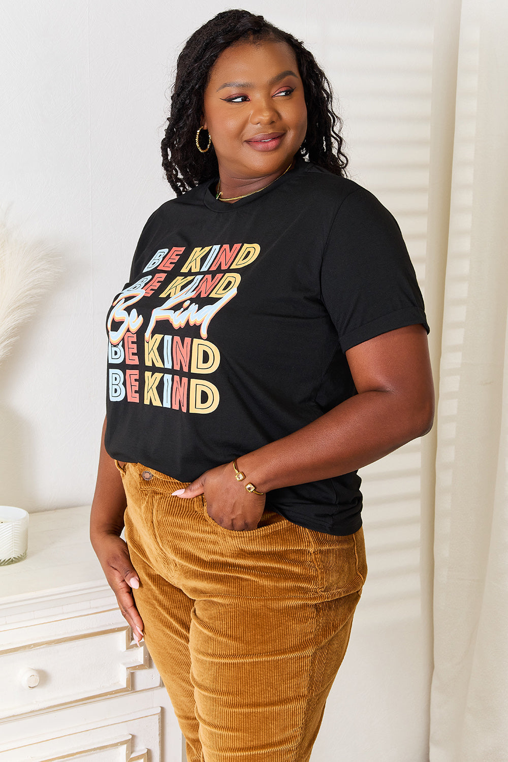 A woman stands with her hands in the pockets of her brown corduroy pants, wearing a casual Simply Love BE KIND Graphic Round Neck T-Shirt that reads "BE KIND" in large colorful letters, showcasing positive message clothing.