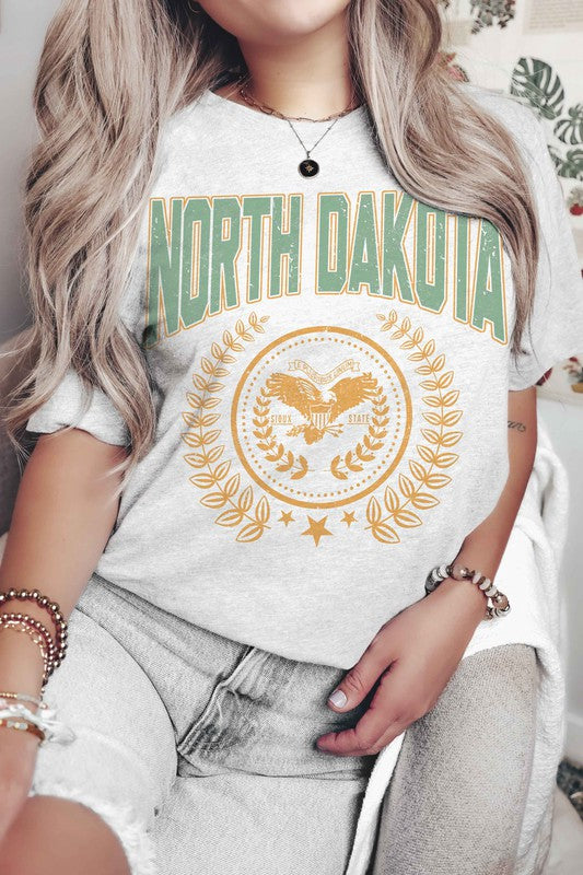 Person wearing a "NORTH DAKOTA Graphic Tee" featuring green and yellow text and graphics, holding a small object in their right hand.