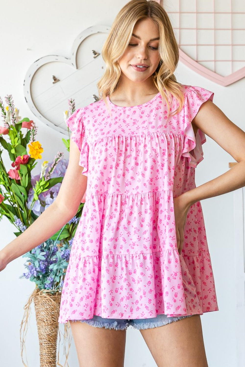 A person wearing the Heimish Full Size Floral Ruffled Tiered Top in pink paired with denim shorts is smiling while standing near a window adorned with vines, radiating a feminine and romantic vibe.