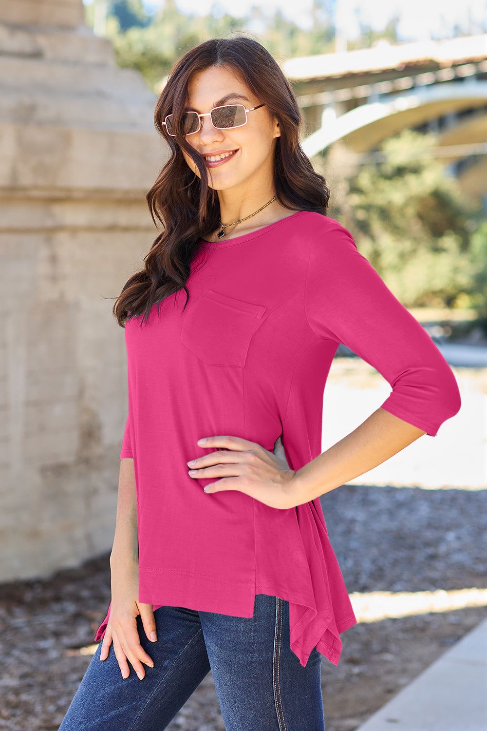 A woman stands outdoors wearing sunglasses, a Basic Bae Full Size Round Neck Pocketed T-Shirt with an asymmetrical hem and chest pocket made of rayon spandex, and blue jeans. She is smiling and has one hand in her pocket. Her basic style radiates effortless charm.