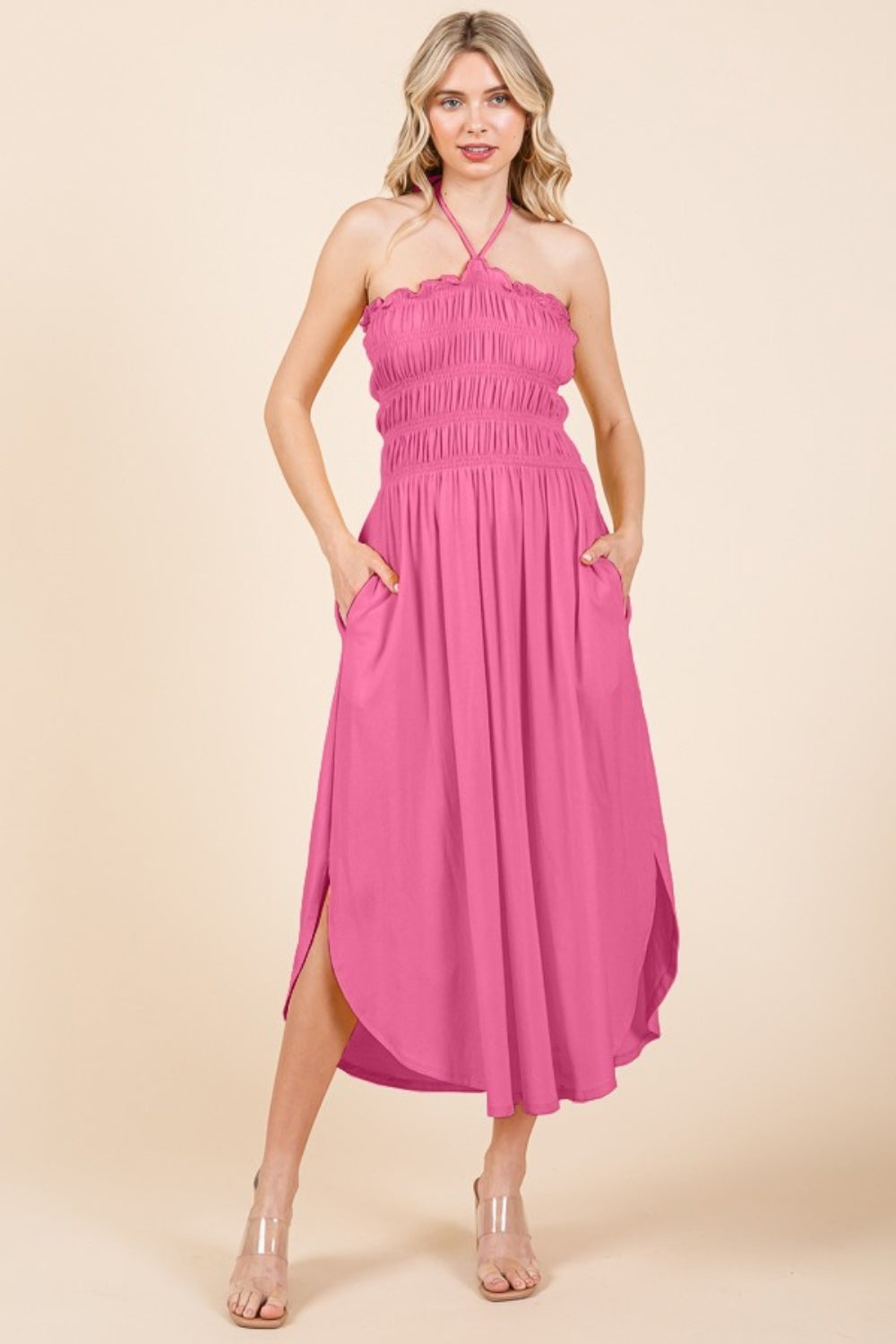 Someone wearing a Culture Code Tie Back Shirring Dress with pockets, featuring a sleeveless and smocked pink halter style, standing against a beige background.