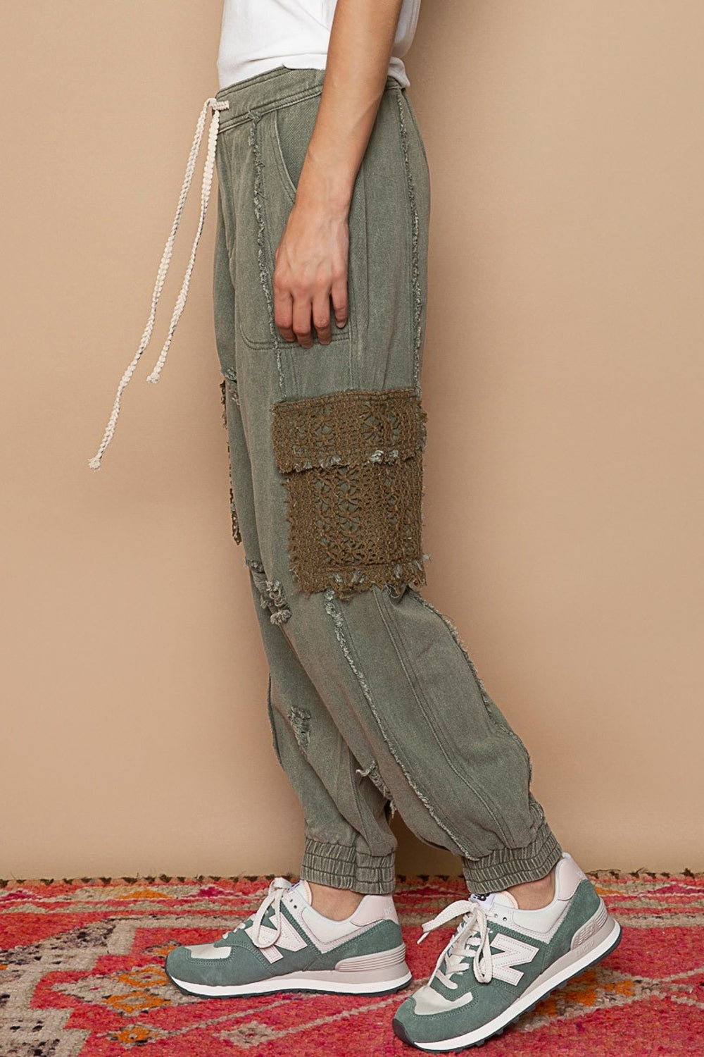 A person sporting POL Distressed Cargo Denim Joggers with Crochet Pockets in green and a white top stands casually on a colorful rug, hands tucked in pockets.