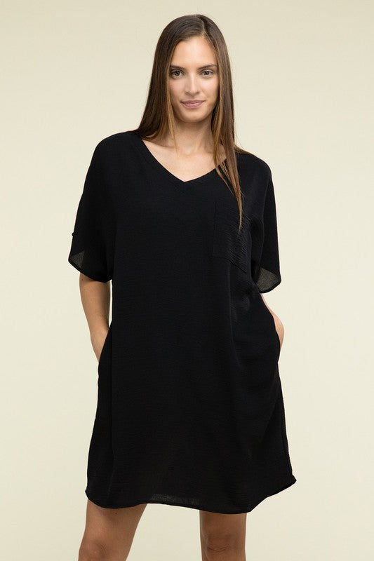 A woman wearing a loose light brown Woven Airflow V Neck T-Shirt Dress with Pockets stands against a plain background. Made from breathable fabric, this summer dress includes pockets where her hands rest as she smiles slightly.