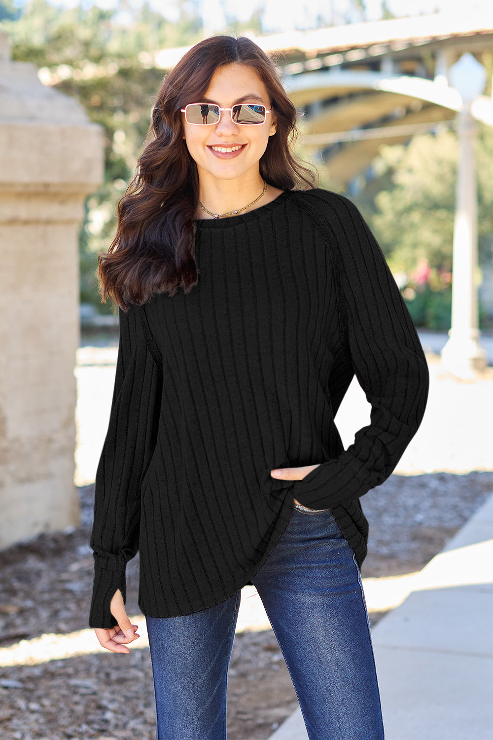 A person with long dark hair and sunglasses stands outdoors, wearing the Basic Bae Full Size Ribbed Round Neck Long Sleeve Knit Top in blue and a pair of jeans. The basic style outfit blends seamlessly with the background of trees and a bridge.