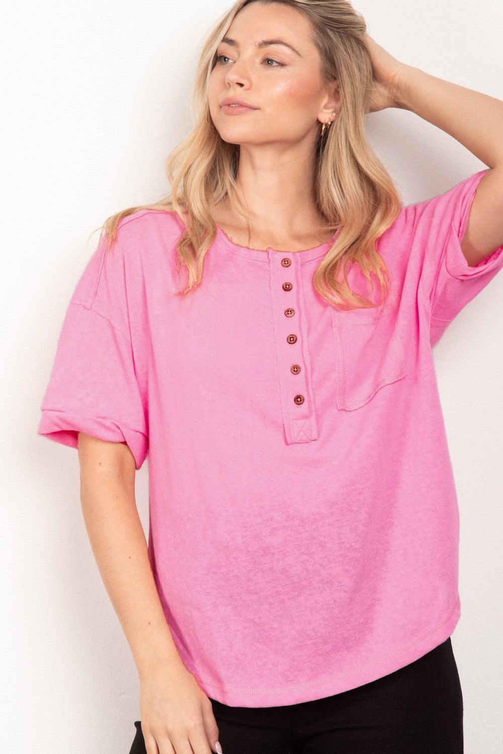 A person with long blonde hair is wearing the VERY J Twisted Sleeve Band Half Button Top in pink, standing in front of a plain white background.