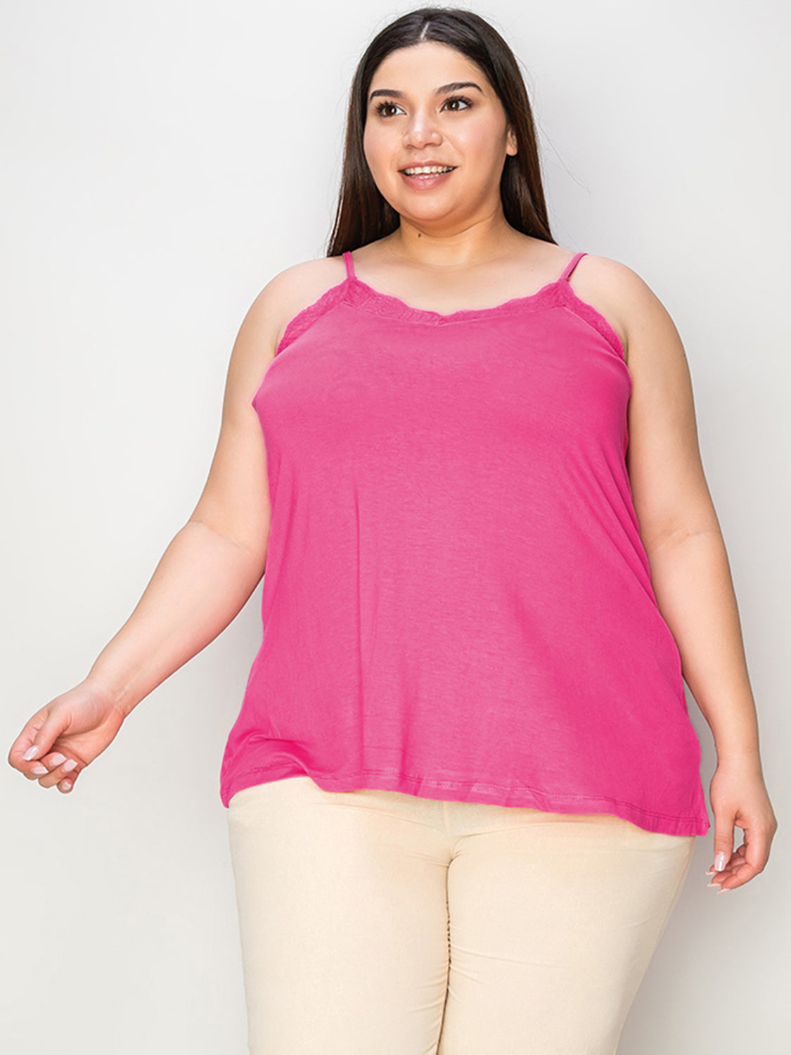 A woman with long hair wearing sunglasses, a sleeveless orange Basic Bae Full Size Lace Trim V-Neck Cami, and blue jeans stands against a concrete wall. She is looking to the side and holding one arm up, touching her sunglasses. Her outfit, available in plus sizes and made from a comfortable rayon blend, perfectly complements her effortless style.