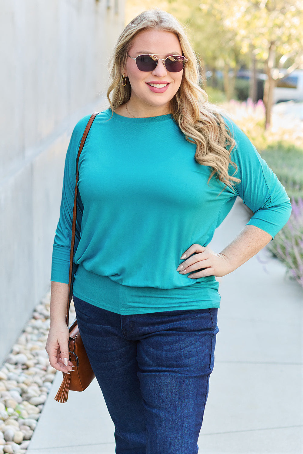 A woman in a slightly stretchy, brown Basic Bae Full Size Round Neck Batwing Sleeve top and blue jeans stands against a concrete wall, holding a brown handbag.