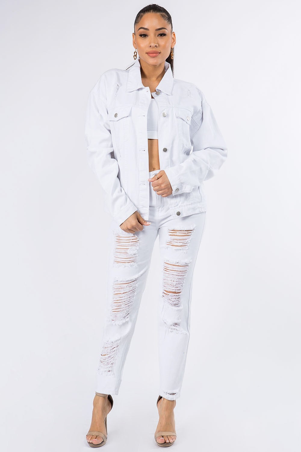 A woman wearing the American Bazi Graphic Distressed Long Sleeve Denim Jacket and matching jeans stands against a plain backdrop. She has her hair styled in a high ponytail and is accessorized with hoop earrings.