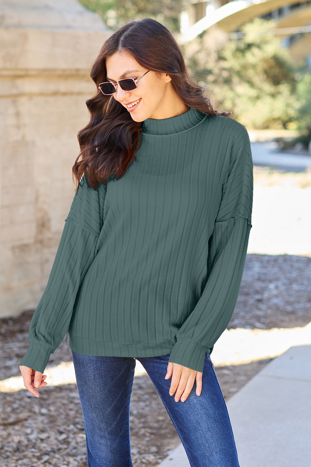 A woman with long hair is wearing a green Basic Bae Full Size Ribbed Exposed Seam Mock Neck Knit Top, blue jeans, and sunglasses. She is standing outdoors and smiling.