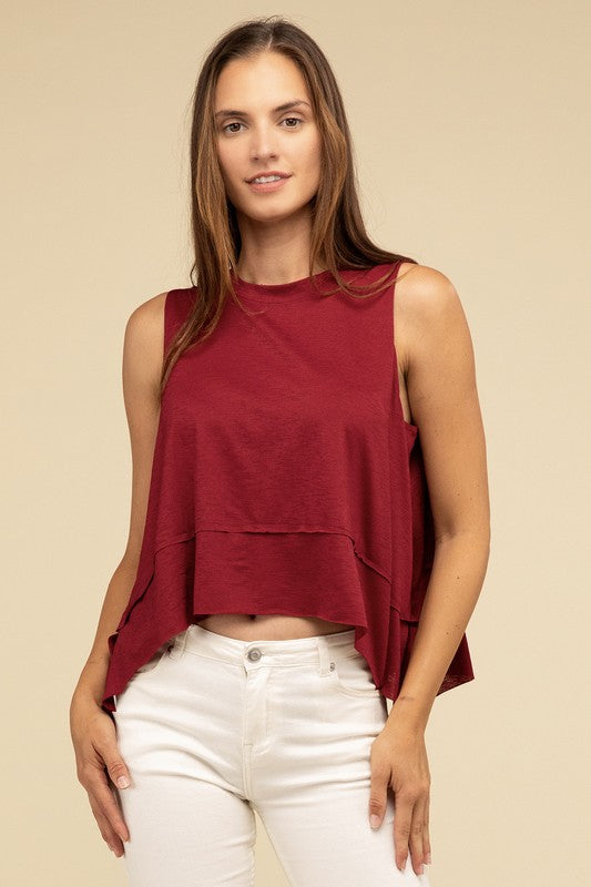 Wearing the Shark Bite Side Slit Short Sleeveless Top in maroon paired with white pants, a model is set against a beige background, exemplifying an effortlessly chic and casual wardrobe.