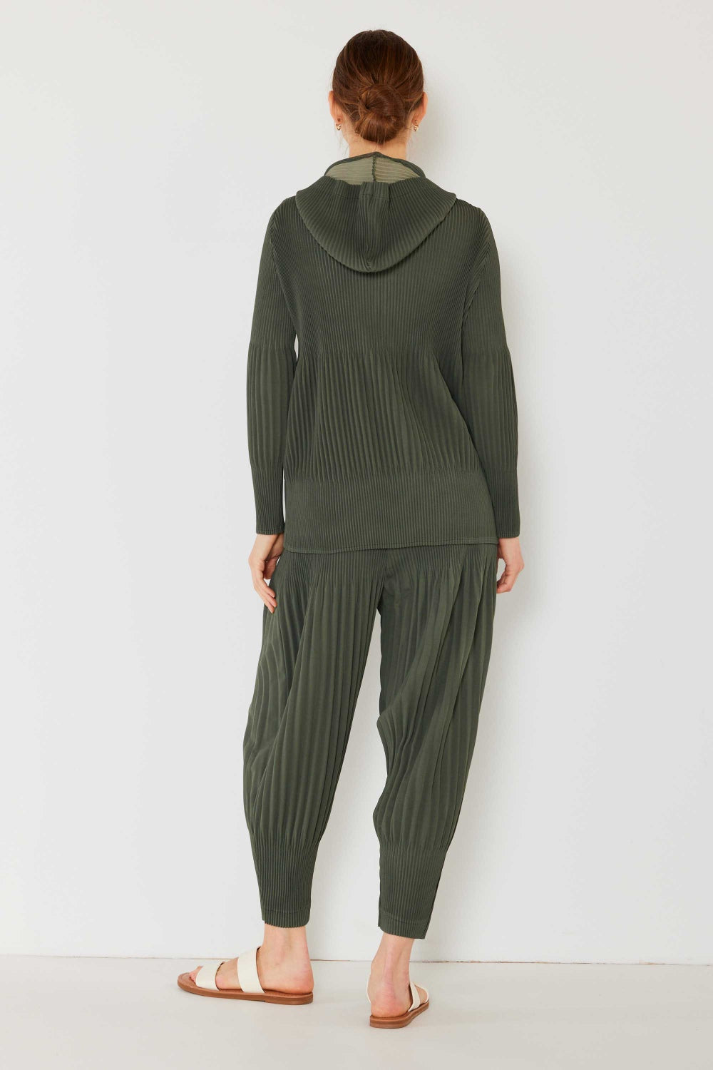 A person stands against a plain background, wearing a matching dark green hooded top and Marina West Swim Pleated Unisex Aladdin Plisse Pants with a ribbed texture, complemented by white sandals.