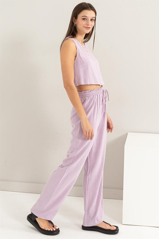 A person is seated on the floor, dressed in a soft lavender D-Linen Blended Top and Pants Set, complemented by chic black sandals.