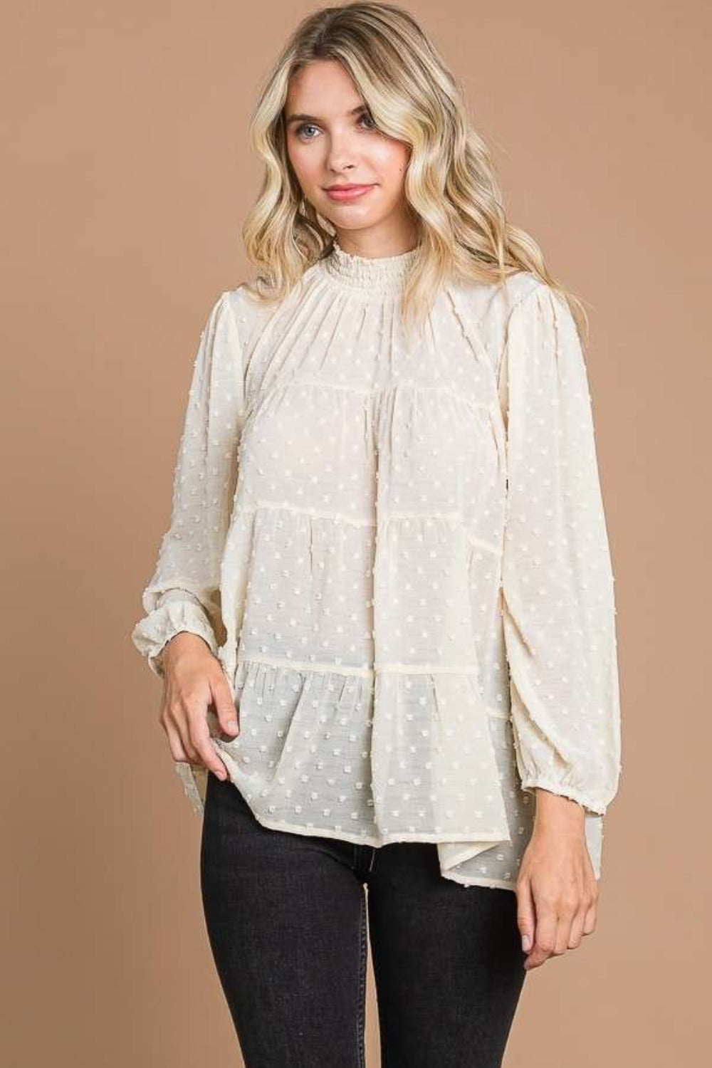 A woman with blonde hair stands against a plain brown background, exuding feminine fashion elegance in a loose, long-sleeved Culture Code Full Size Swiss Dot Smocked Mock Neck Blouse.