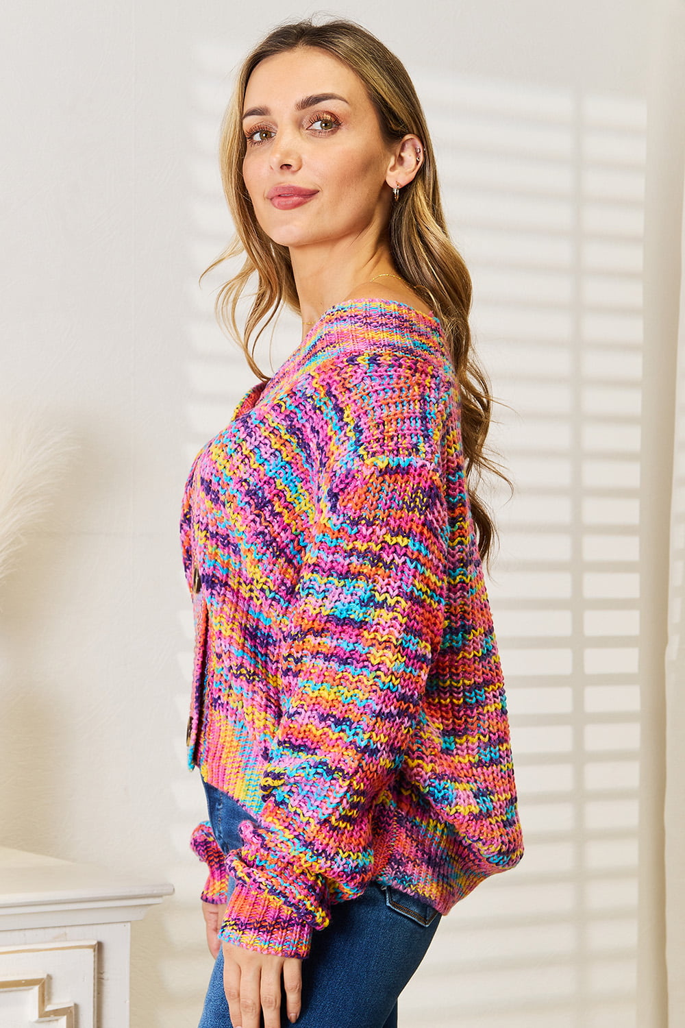 Person wearing an Angel Wings Woven Right V-Neck Long Sleeve Cardigan, characterized by its multicolored design and large buttons, paired with blue jeans. The cardigan, made of 100% acrylic knit, offers a versatile look perfect for any occasion. They are standing indoors against a white background.