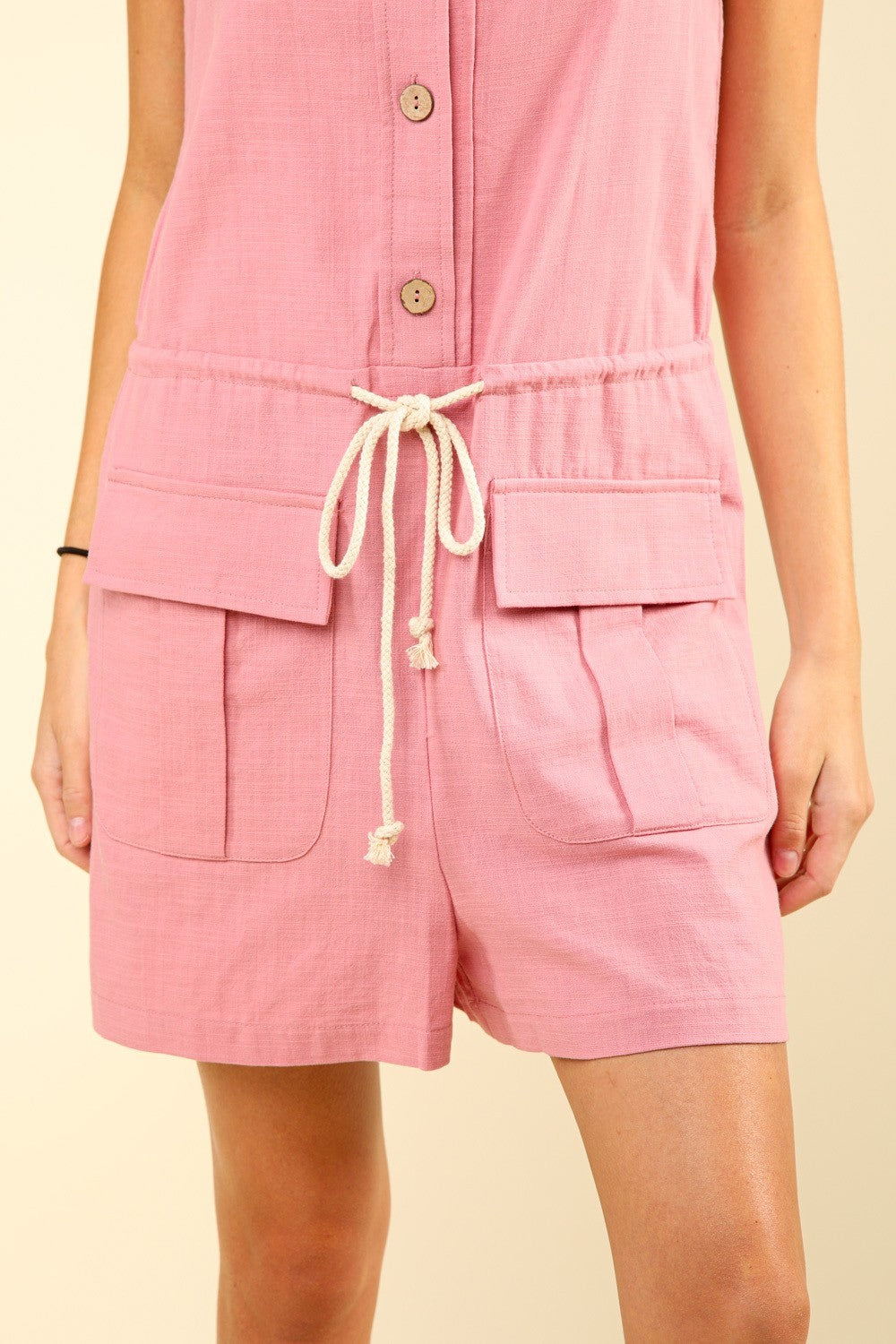 A woman wearing the VERY J Half Button Drawstring Sleeveless Romper, styled in pink, poses against a neutral backdrop.