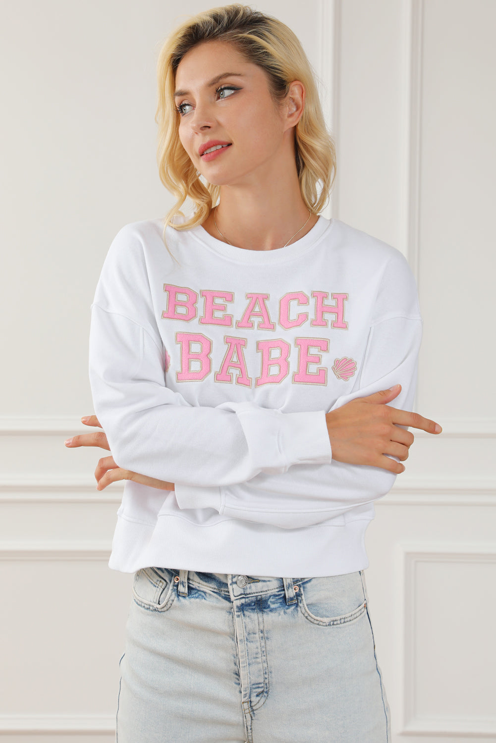 A person with long blonde hair is standing with their back to the camera, wearing a White BEACH BABE Slogan Graphic Casual Sweatshirt and light blue jeans.