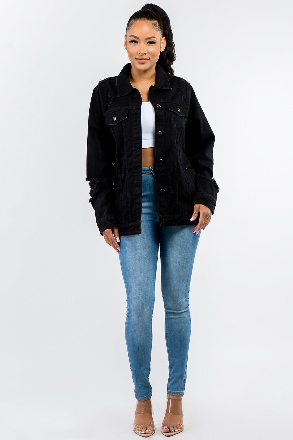 A woman stands against a plain background, epitomizing casual outfits with a touch of vintage look, wearing the American Bazi Full Size Button Up Distressed Denim Jacket over a white crop top and jeans.