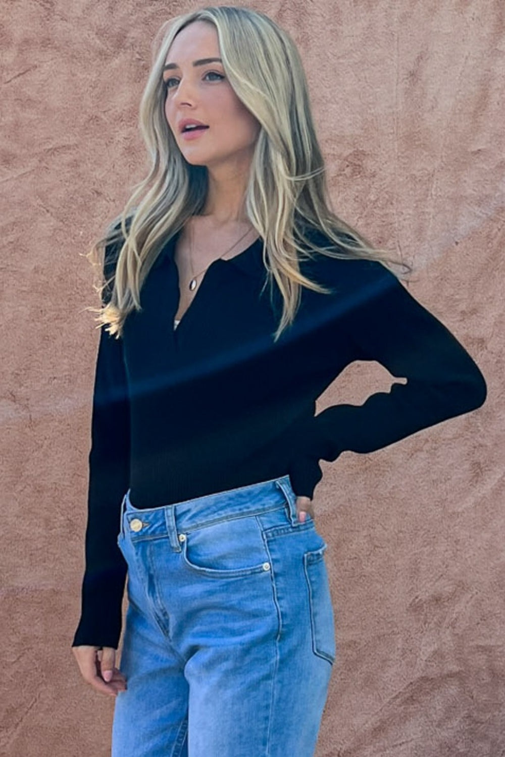 A woman with long blonde hair is standing against a ribbed-texture wall, wearing the And The Why Full Size Johnny Collar Long Sleeve Ribbed Bodysuit in black and blue jeans, with one hand in her pocket.