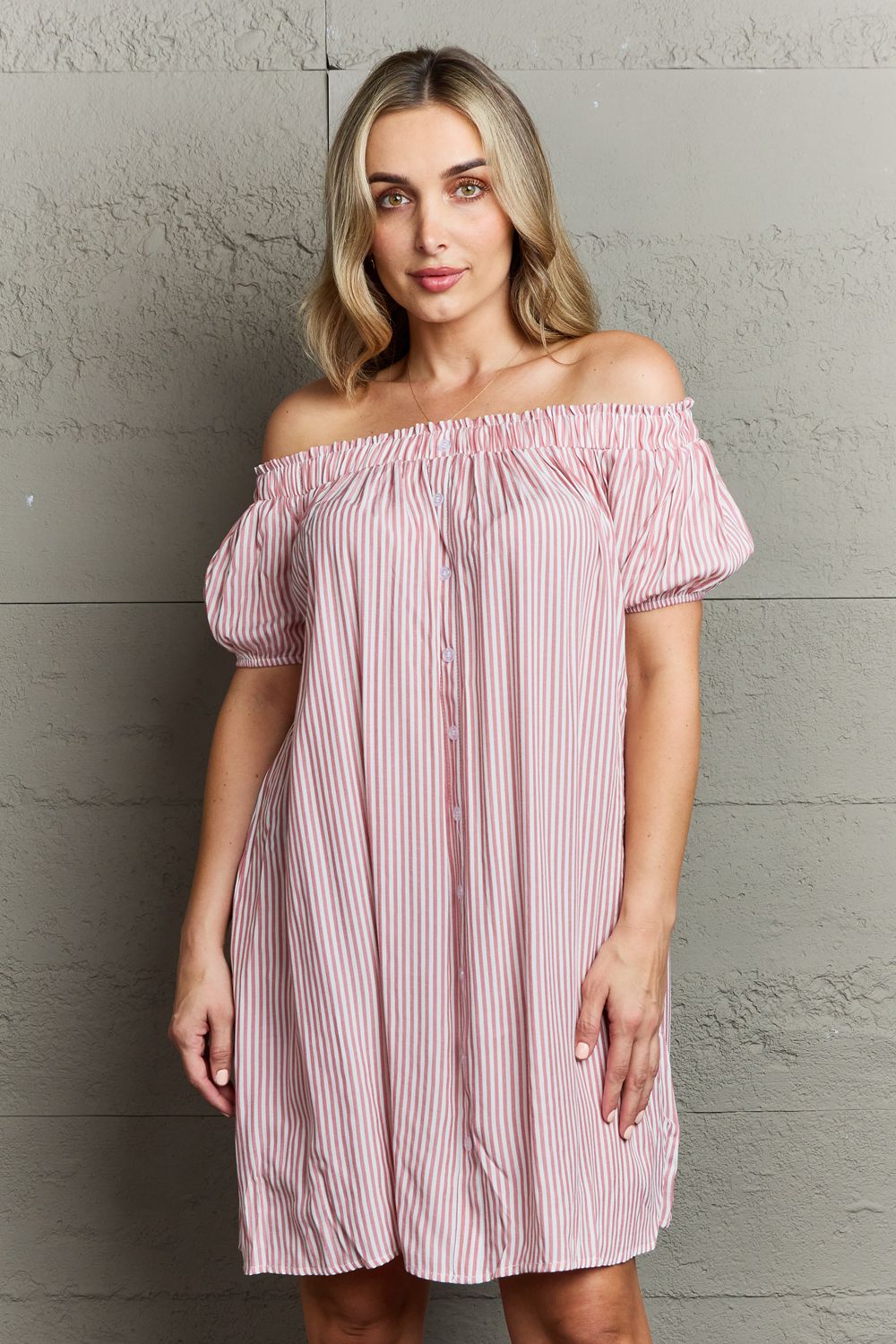 A woman exudes sophisticated elegance in the Ninexis Show Compassion Off The Shoulder Mini Dress, a pink and white striped ensemble, as she stands against a gray textured wall.