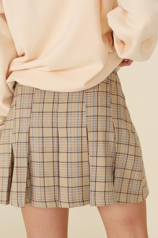 A person dressed in a plaid pleated mini skirt paired with a beige sweater is posed against a plain backdrop. This fashionable and versatile outfit, featuring the Plaid Pleated Mini Skirt, is designed for easy maintenance and can be machine washed in cold water.