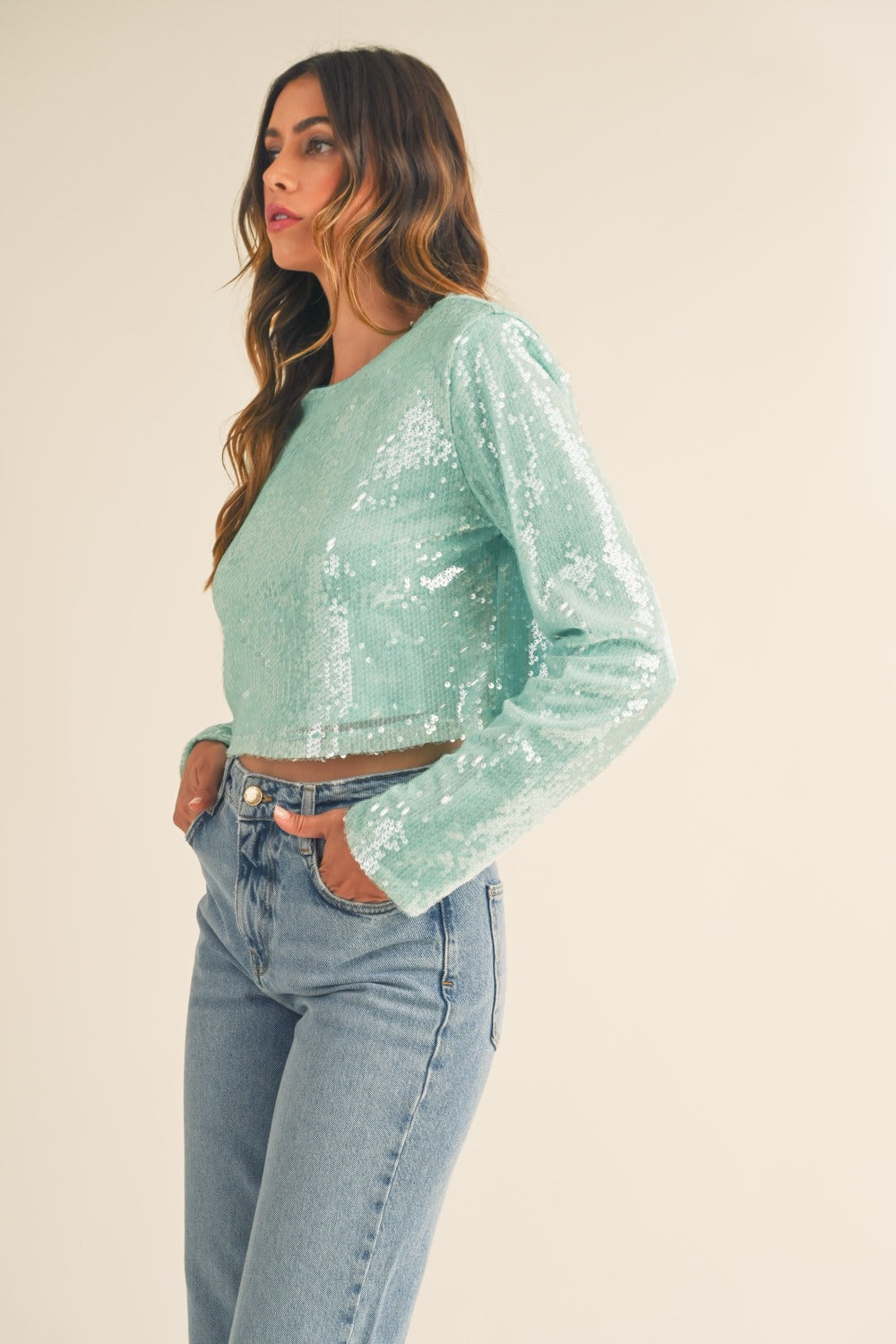 A person with long hair, wearing a fashion-forward, seafoam green MABLE Shoulder Padded Sequin Crop Top and high-waisted blue jeans, stands against a plain background.