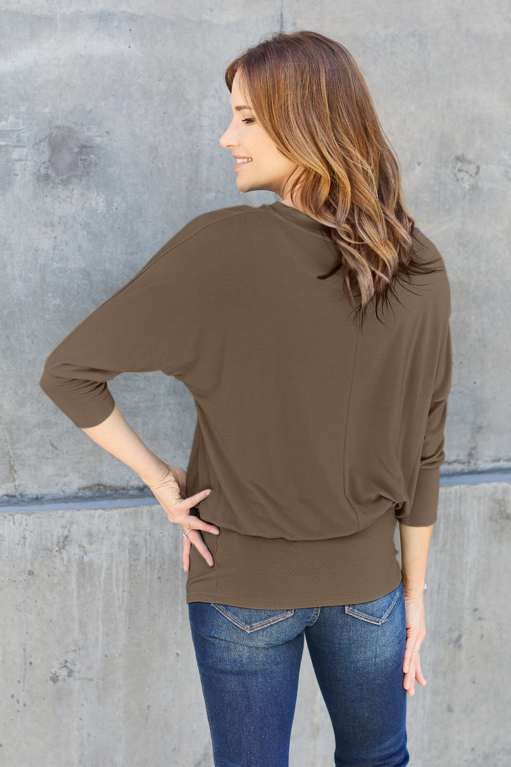 A woman in a slightly stretchy, brown Basic Bae Full Size Round Neck Batwing Sleeve top and blue jeans stands against a concrete wall, holding a brown handbag.