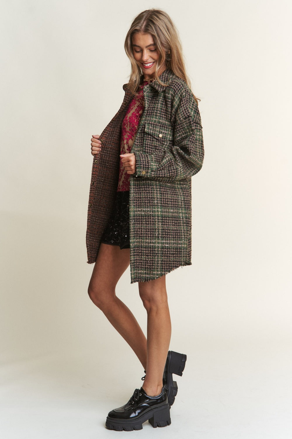 A woman wearing an eclectic mix poses against a plain background, showcasing an ideal fall wardrobe ensemble. She dons a mixed-patterned oversized jacket over a red floral sheer top and black sequined shorts. The J.NNA Snap Down Tweed Plaid Contrast Longline Shacket adds texture and depth to her look.