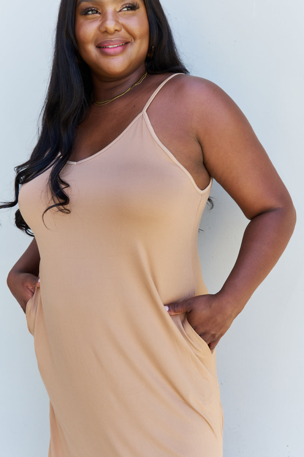 A woman stands against a plain background, wearing the Ninexis Good Energy Full Size Cami Side Slit Maxi Dress in Camel along with a wide-brimmed hat. She has one hand on her hip and is smiling.