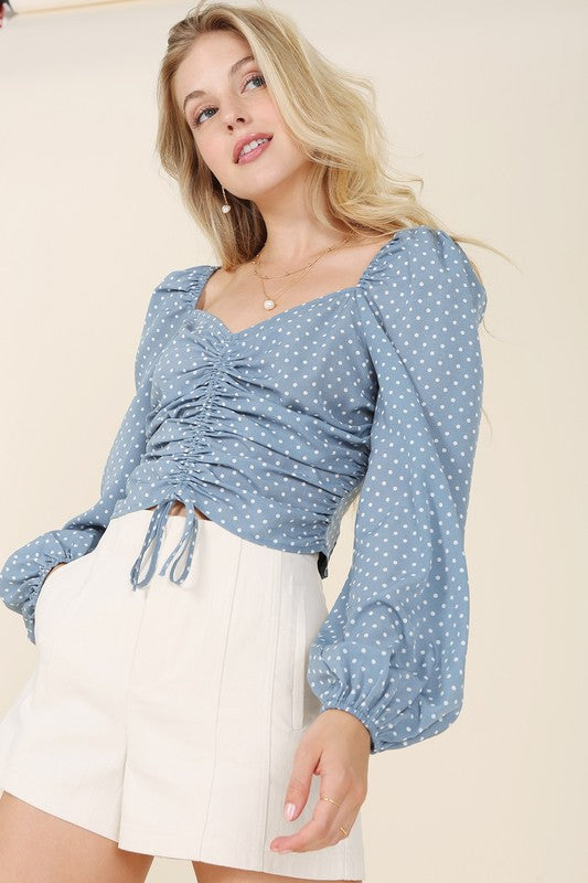 A person wearing the Ruched polka dot crop top with puff sleeves and white shorts poses against a light background.