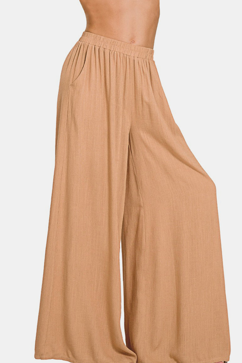 A person wearing Zenana Pleated Linen Blend Wide Leg Pants in tan, with hands in pockets, standing against a plain white background. Upper body and face are not visible.