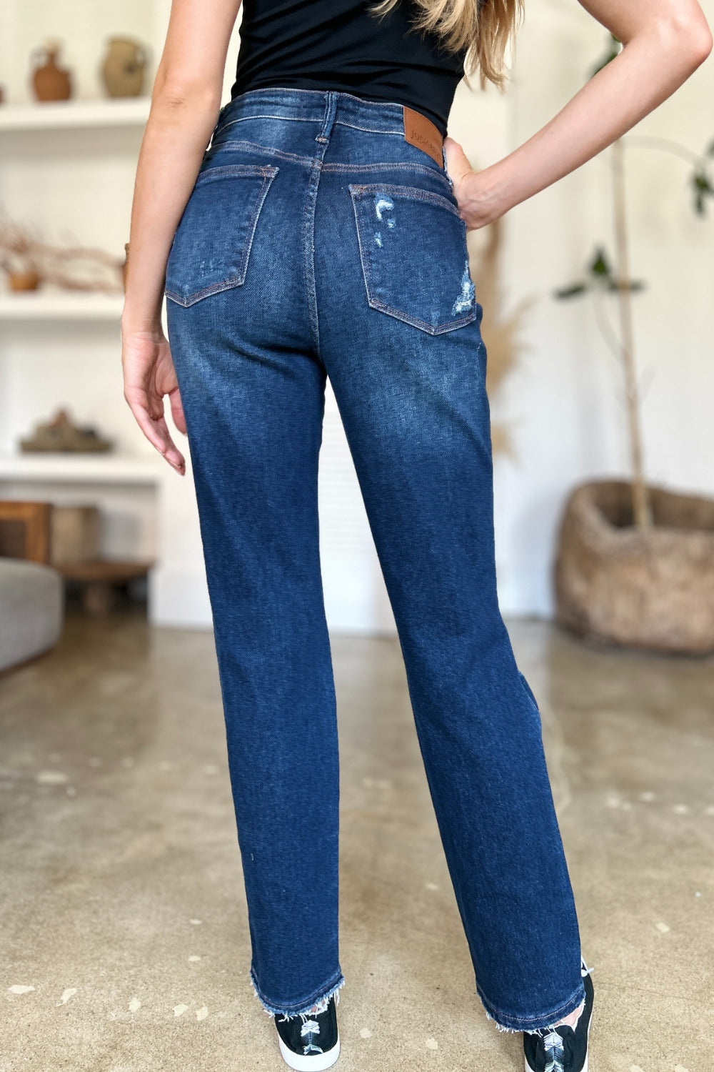 A woman in a black sleeveless top and Judy Blue Full Size High Waist Rigid Magic Heavy Destroy Straight Jeans stands indoors with her hands in her pockets. The background features a plant and shelves adorned with decor items.