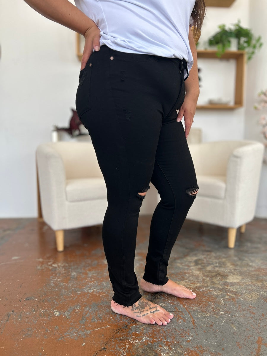 A person wearing a white T-shirt and Judy Blue Full Size Distressed Tummy Control High Waist Skinny Jeans poses barefoot in a room with white chairs and decorative plants in the background.