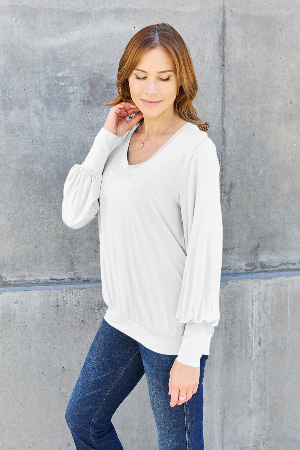 A woman with brown hair is standing against a concrete wall, wearing the Basic Bae Full Size V-Neck Lantern Sleeve Top in teal and blue basic-style jeans, smiling.