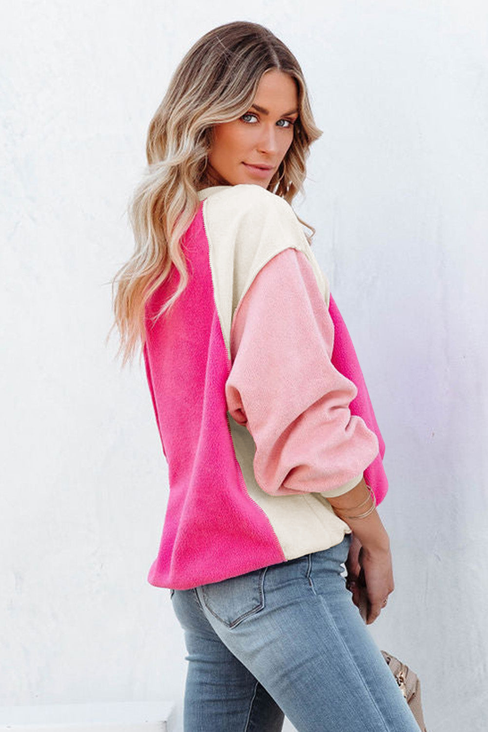 A person with long blonde hair is standing facing away from the camera against a beige background, showcasing the comfortable fleece fabric of the Rose Colorblock Long Sleeve Pullover Fleece Sweatshirt in pink shades and light blue jeans.