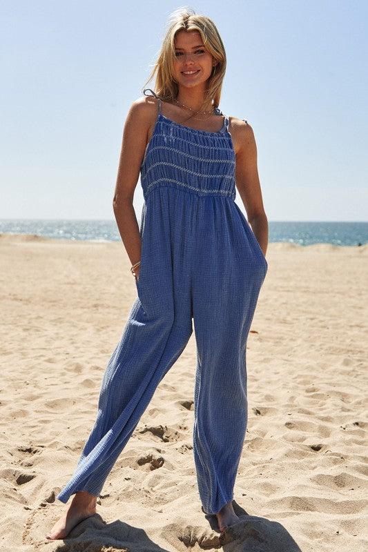 A person wearing the Washed Multi Smocked Detail Tie Straps Jumpsuit stands on the sunny beach, with the ocean creating a perfect backdrop.