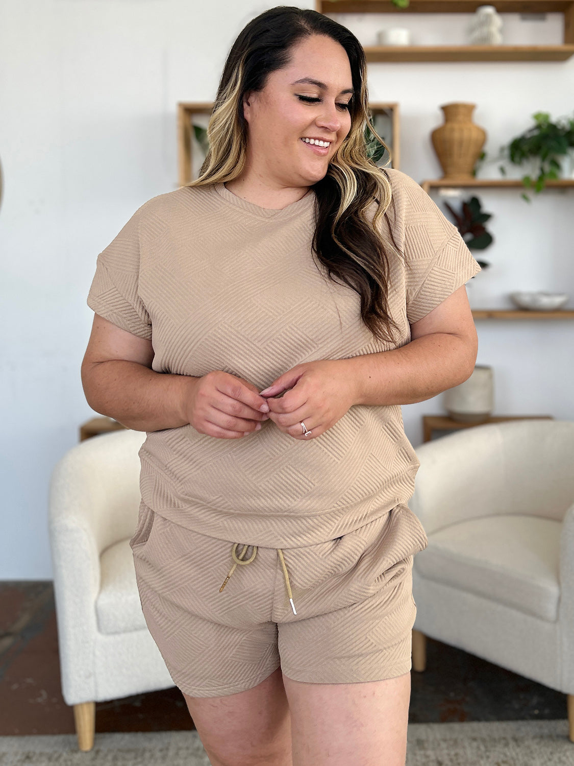 A woman with long dark hair is wearing the Double Take Full Size Texture Short Sleeve T-Shirt and Drawstring Shorts Set, made from beige polyester. The set features a short-sleeve top and matching shorts, with the woman standing confidently with one hand in her pocket and maintaining a neutral expression. For easy care, remember to machine wash cold.