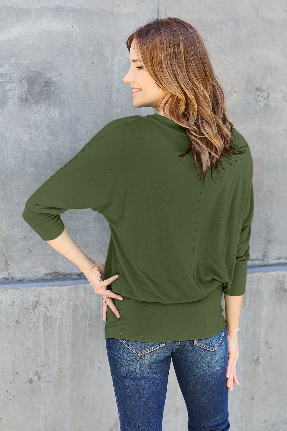 A woman in a slightly stretchy, brown Basic Bae Full Size Round Neck Batwing Sleeve top and blue jeans stands against a concrete wall, holding a brown handbag.