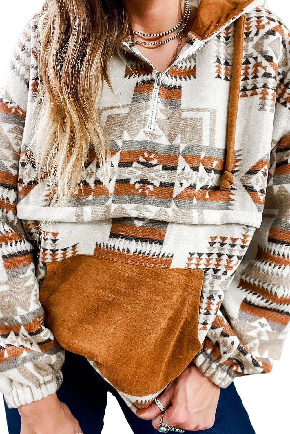 A person with long blonde hair, seen from behind, is wearing the Beige Aztec Print Kangaroo Pocket Half-Zip Hoodie. The hoodie showcases an Aztec pattern in shades of brown, beige, and orange and features a drop shoulder design with a cozy kangaroo pocket, all while standing outdoors.