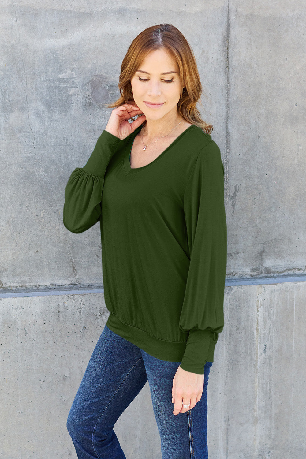 A woman with brown hair is standing against a concrete wall, wearing the Basic Bae Full Size V-Neck Lantern Sleeve Top in teal and blue basic-style jeans, smiling.