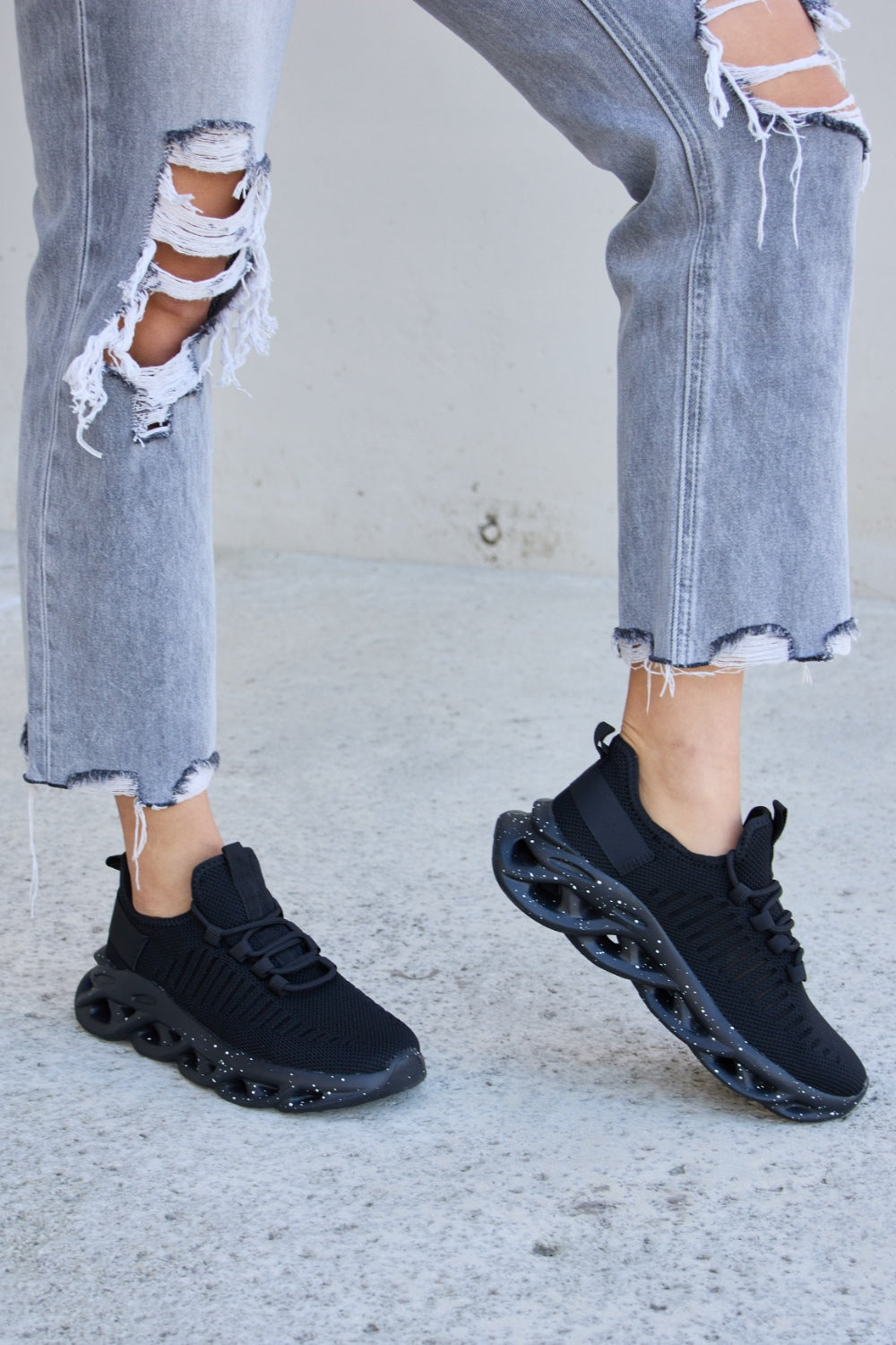 A person wearing ripped gray jeans and Forever Link Mesh Breathable Lace-Up Athletic Shoes is standing on a concrete surface.