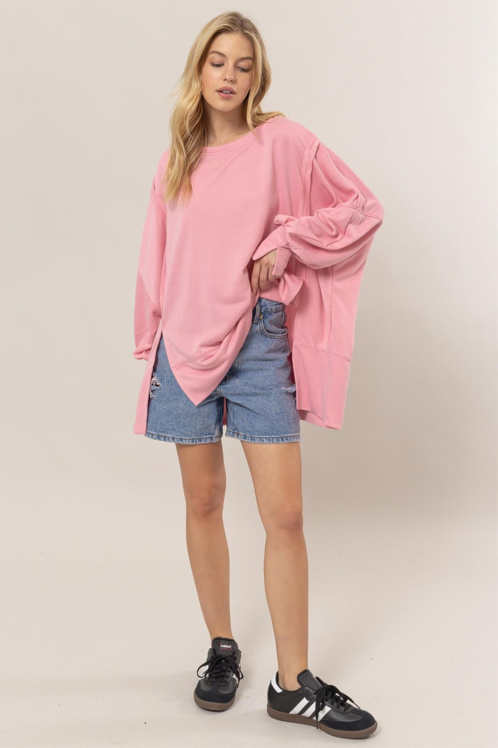 A person with long hair wearing a comfortable, stylish HYFVE French Terry Long Sleeve High-Low Slit Sweatshirt in pink, denim shorts, and black athletic shoes stands against a plain backdrop.