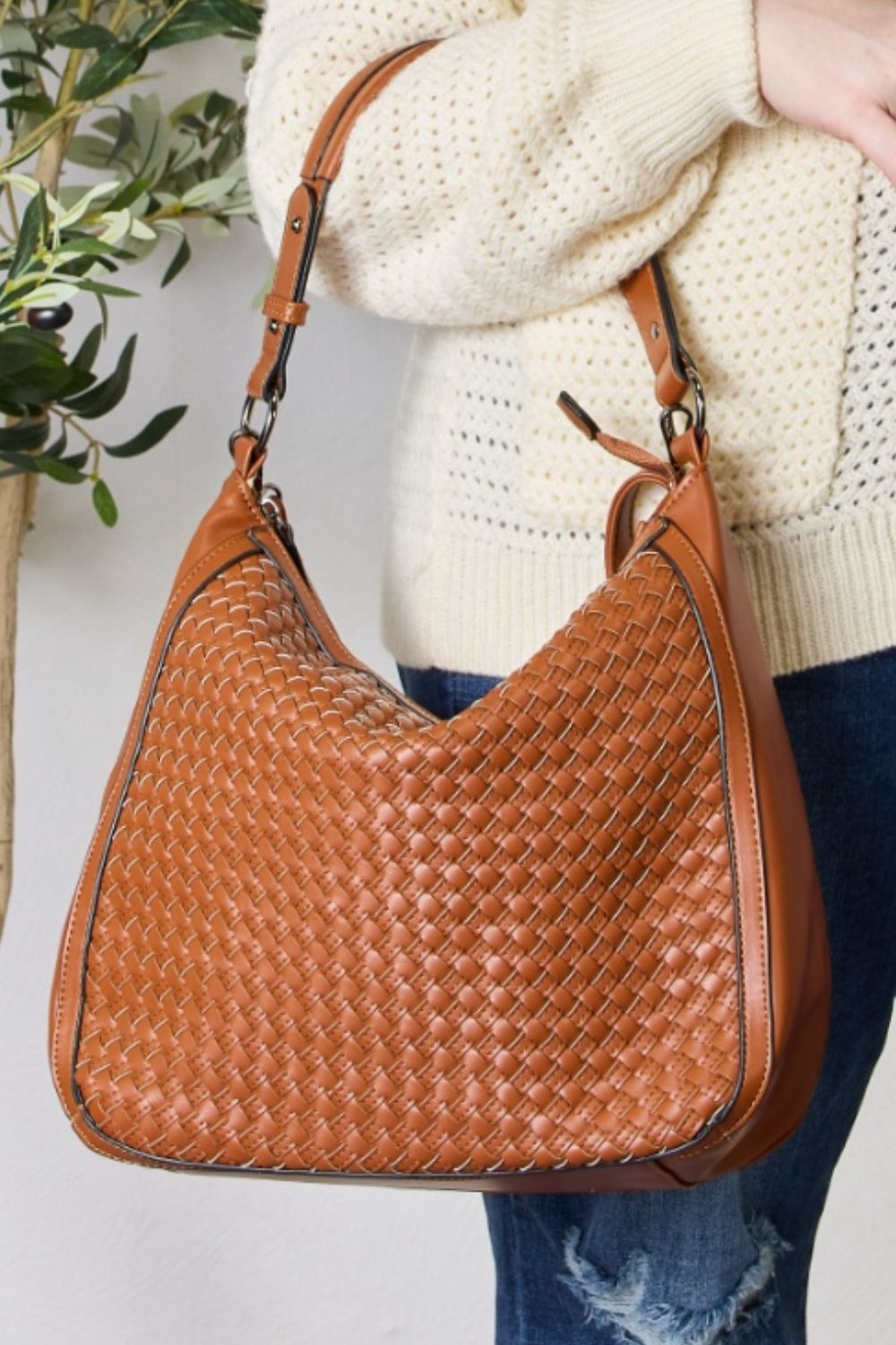 A person carries the SHOMICO Weaved Vegan Leather Handbag in a stylish brown hue, featuring a sleek crossbody strap, expertly paired with a cream sweater and jeans.