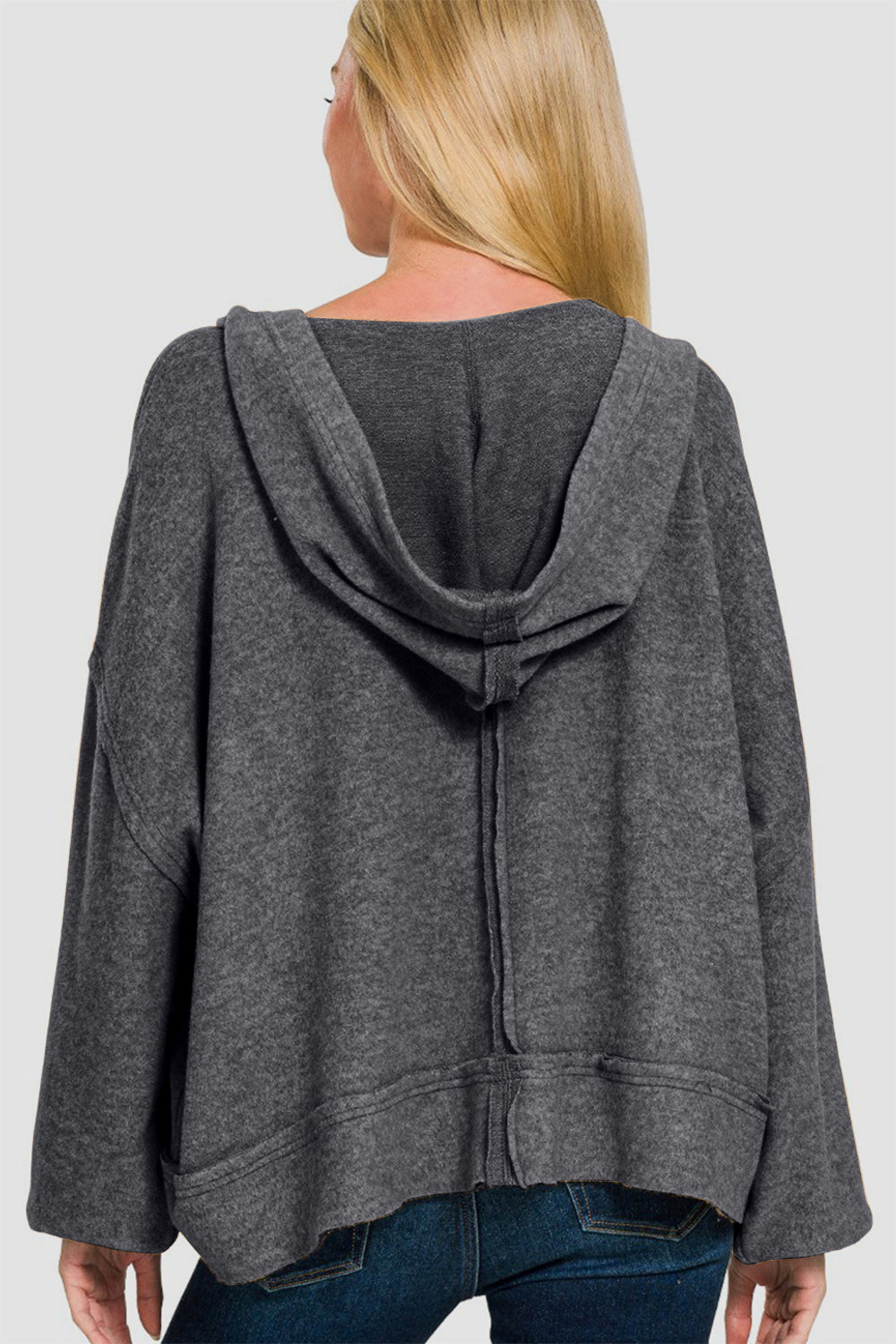 A person with long blond hair stands against a white background, wearing the Zenana Brushed Hacci Exposed Seam Hoodie in dark gray paired with jeans.